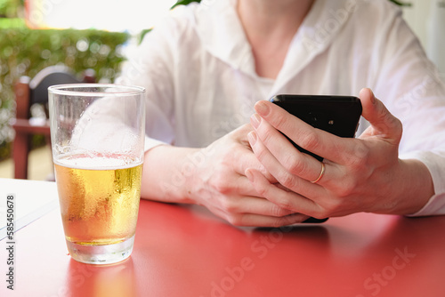 woman at a street cafe table holds a smartphone in her hands. There is a glass of beer on the table. Day break, surfing the internet, meeting up with friends.