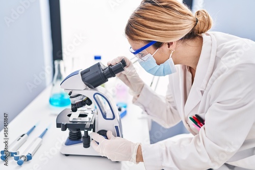 Young blonde woman scientist wearing medical mask using microscope at laboratory