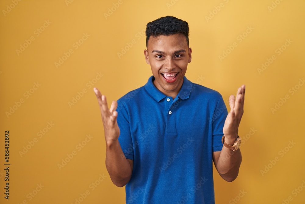 Young hispanic man standing over yellow background celebrating mad and crazy for success with arms raised and closed eyes screaming excited. winner concept