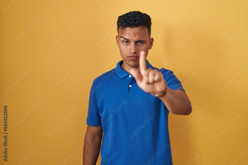 Young hispanic man standing over yellow background pointing with finger up and angry expression, showing no gesture Young hispanic man standing over yellow background pointing with finger up and angry expression, showing no gesture