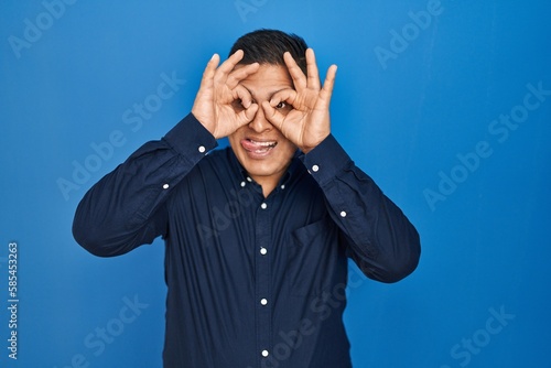 Hispanic young man standing over blue background doing ok gesture like binoculars sticking tongue out, eyes looking through fingers. crazy expression.