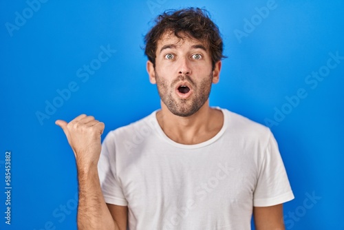 Hispanic young man standing over blue background surprised pointing with hand finger to the side, open mouth amazed expression.