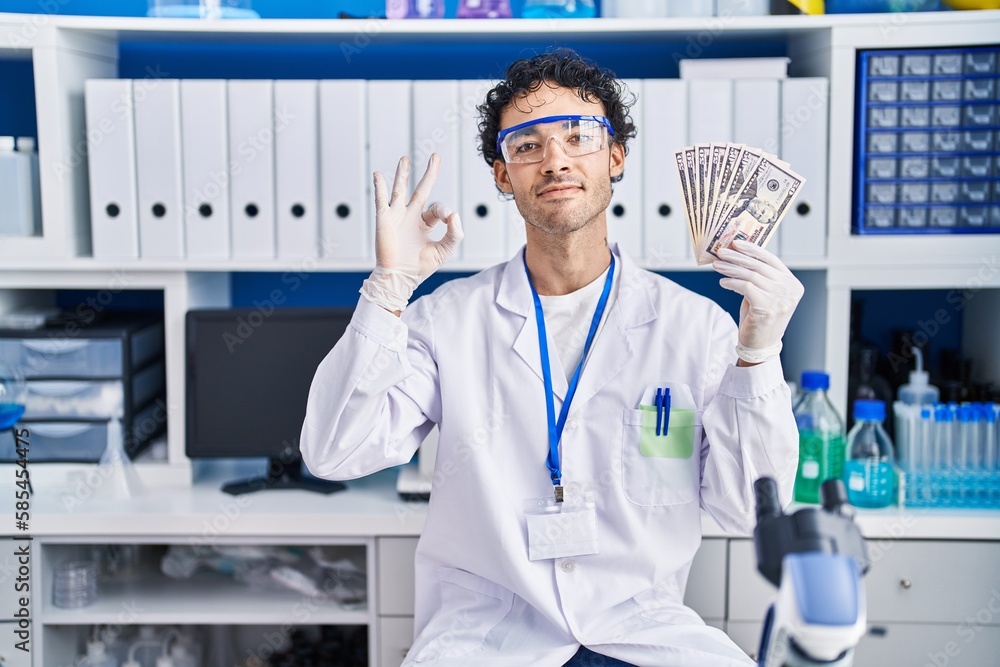 Hispanic man working at scientist laboratory holding money doing ok sign with fingers, smiling friendly gesturing excellent symbol Hispanic man working at scientist laboratory holding money doing ok sign with fingers, smiling friendly gesturing excellent symbol