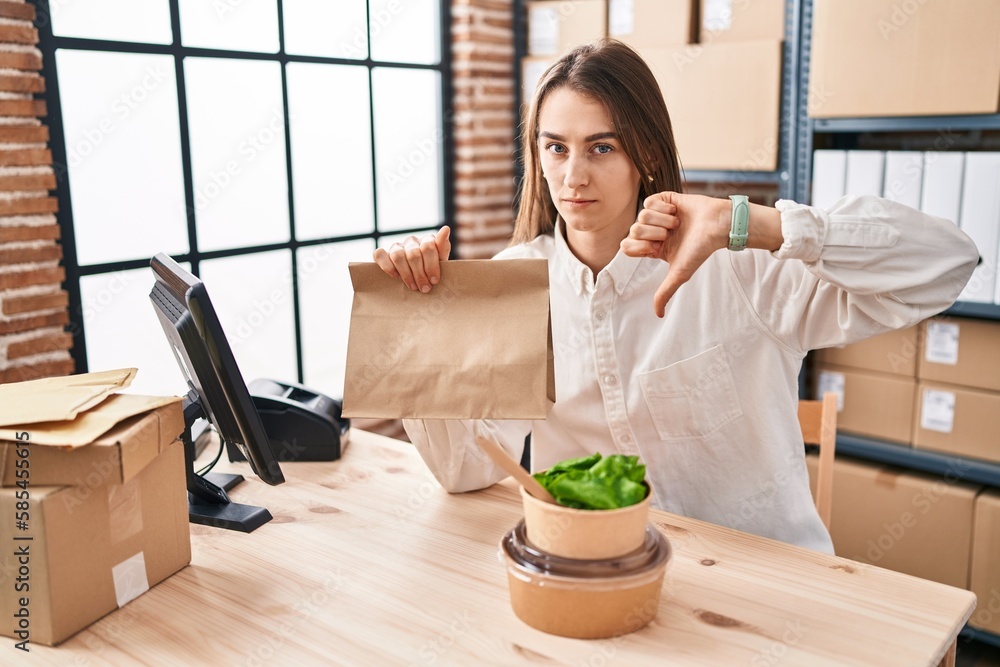 Young caucasian woman working at small business ecommerce holding take away food with angry face, negative sign showing dislike with thumbs down, rejection concept Young caucasian woman working at small business ecommerce holding take away food with angry face, negative sign showing dislike with thumbs down, rejection concept