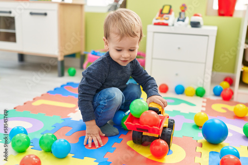 Adorable blond toddler playing with tractor and balls sitting on floor at kindergarten
