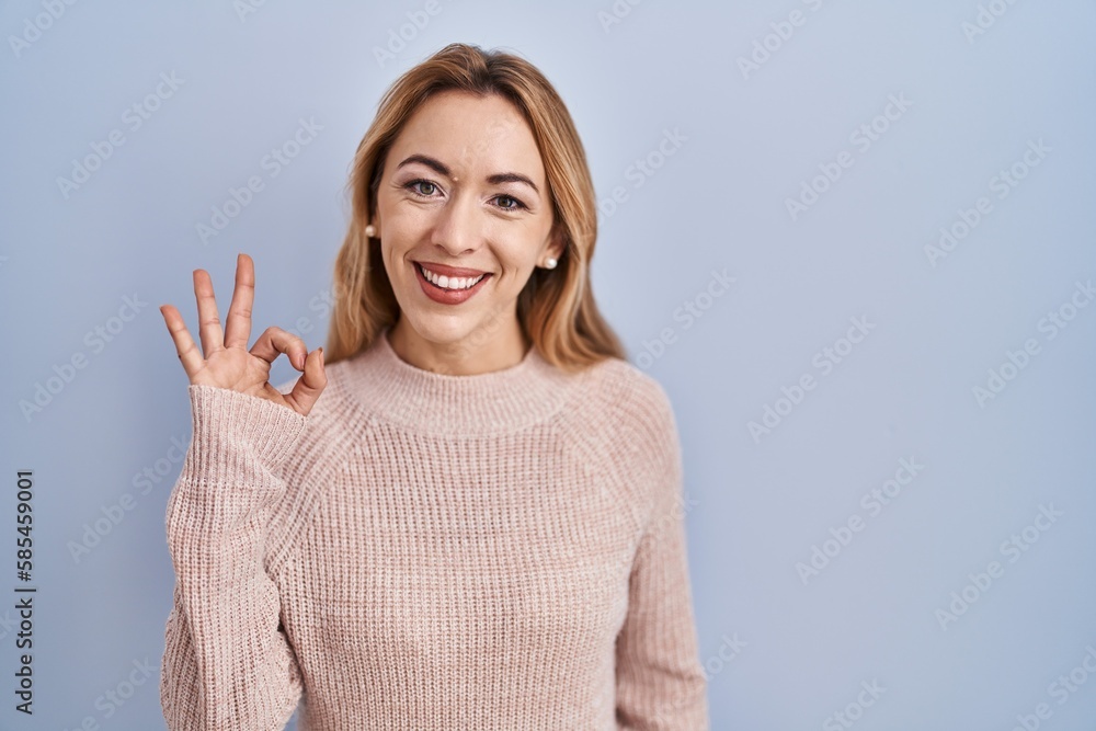 Hispanic woman standing over blue background smiling positive doing ok sign with hand and fingers. successful expression. Hispanic woman standing over blue background smiling positive doing ok sign with hand and fingers. successful expression.