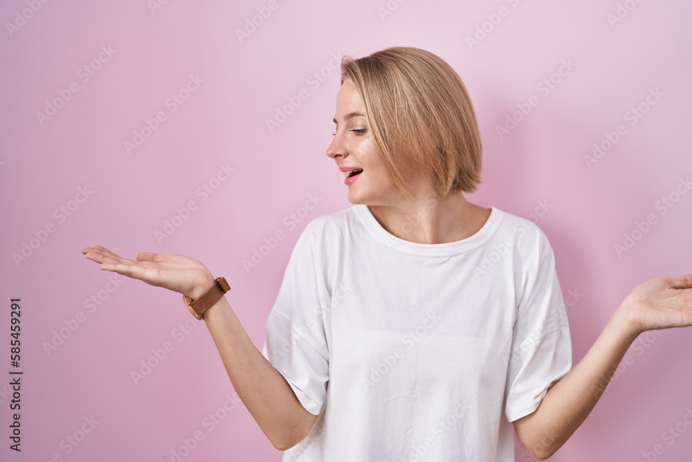 Young caucasian woman standing over pink background smiling showing both hands open palms, presenting and advertising comparison and balance Young caucasian woman standing over pink background smiling showing both hands open palms, presenting and advertising comparison and balance