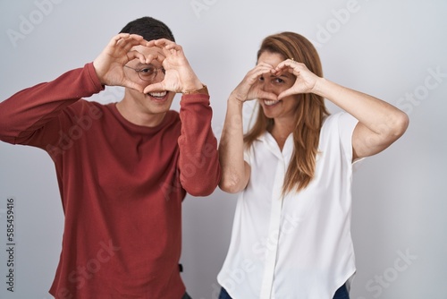 Mother and son standing together over isolated background doing heart shape with hand and fingers smiling looking through sign