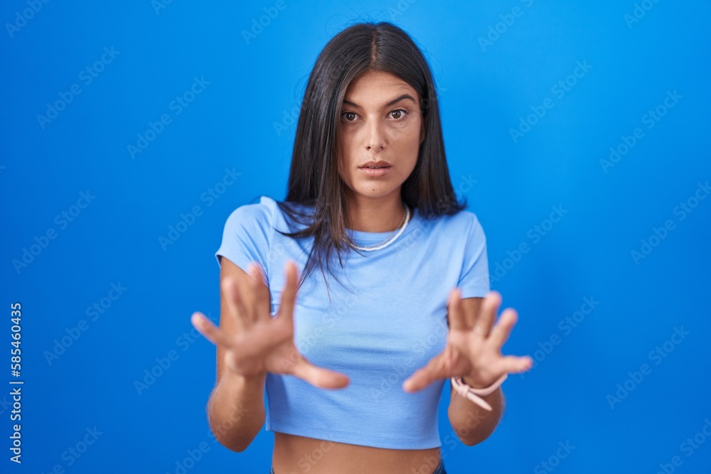 Brunette young woman standing over blue background afraid and terrified with fear expression stop gesture with hands, shouting in shock. panic concept. Brunette young woman standing over blue background afraid and terrified with fear expression stop gesture with hands, shouting in shock. panic concept.