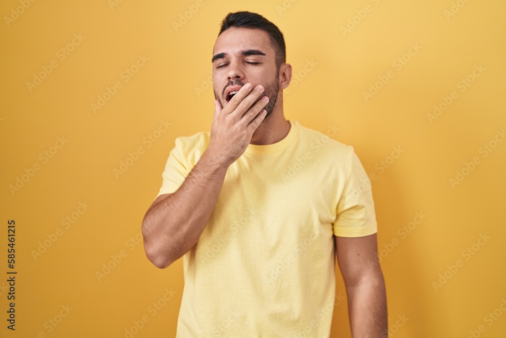 Young hispanic man standing over yellow background bored yawning tired covering mouth with hand. restless and sleepiness. Young hispanic man standing over yellow background bored yawning tired covering mouth with hand. restless and sleepiness.