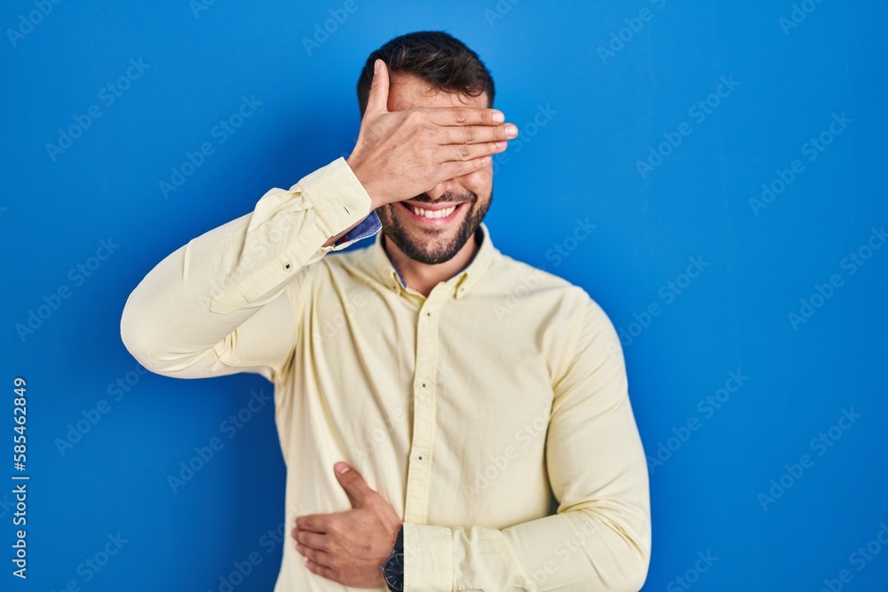 Handsome hispanic man standing over blue background smiling and laughing with hand on face covering eyes for surprise. blind concept. Handsome hispanic man standing over blue background smiling and laughing with hand on face covering eyes for surprise. blind concept.