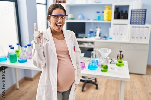 Young pregnant woman working at scientist laboratory pointing finger up with successful idea. exited and happy. number one.