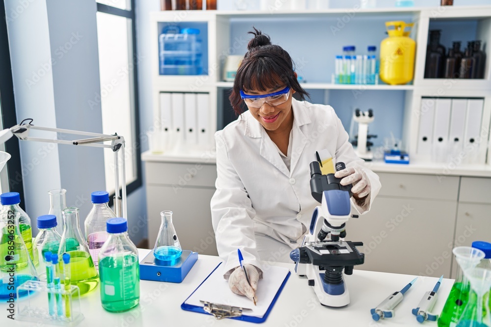 Young beautiful latin woman scientist using microscope writing report at laboratory Young beautiful latin woman scientist using microscope writing report at laboratory