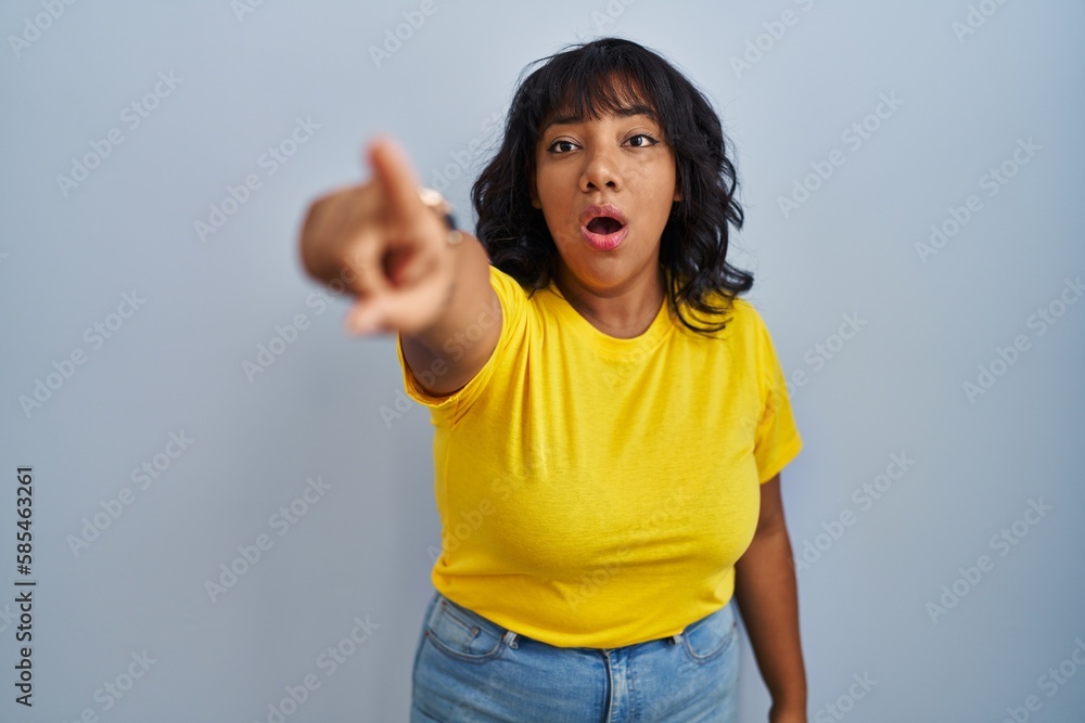 Hispanic woman standing over blue background pointing with finger surprised ahead, open mouth amazed expression, something on the front
