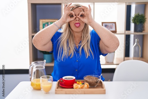 Caucasian plus size woman eating breakfast at home doing ok gesture like binoculars sticking tongue out, eyes looking through fingers. crazy expression.