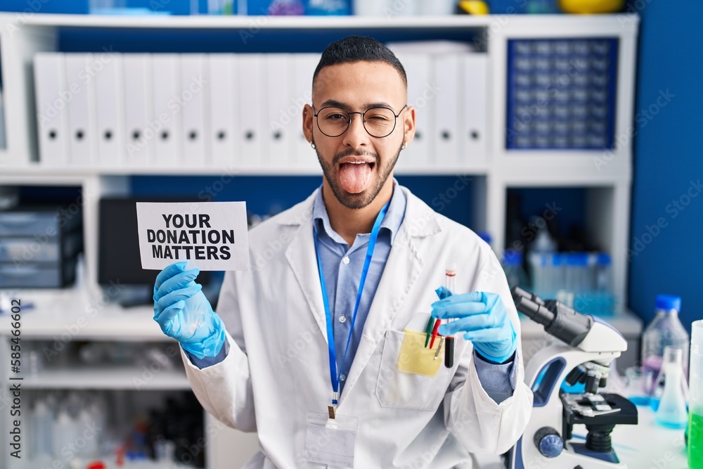 Young hispanic man working at scientist laboratory holding your donation matters holding blood sample sticking tongue out happy with funny expression. Young hispanic man working at scientist laboratory holding your donation matters holding blood sample sticking tongue out happy with funny expression.