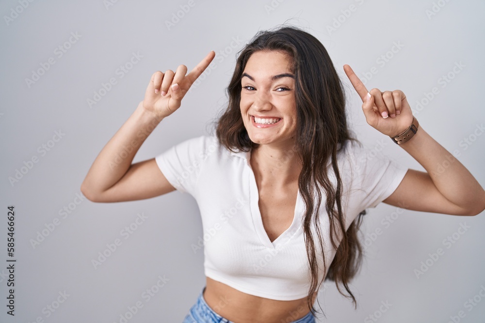 Young teenager girl standing over white background smiling amazed and surprised and pointing up with fingers and raised arms. Young teenager girl standing over white background smiling amazed and surprised and pointing up with fingers and raised arms.