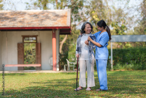 Asian nurse or female caregiver helping senior woman holding cane to walk in the hospital garden.