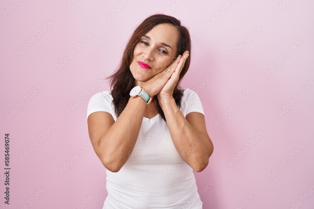 Middle age brunette woman standing over pink background sleeping tired dreaming and posing with hands together while smiling with closed eyes. Middle age brunette woman standing over pink background sleeping tired dreaming and posing with hands together while smiling with closed eyes.