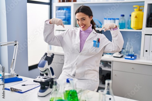 Young brunette woman working at scientist laboratory looking confident with smile on face  pointing oneself with fingers proud and happy.