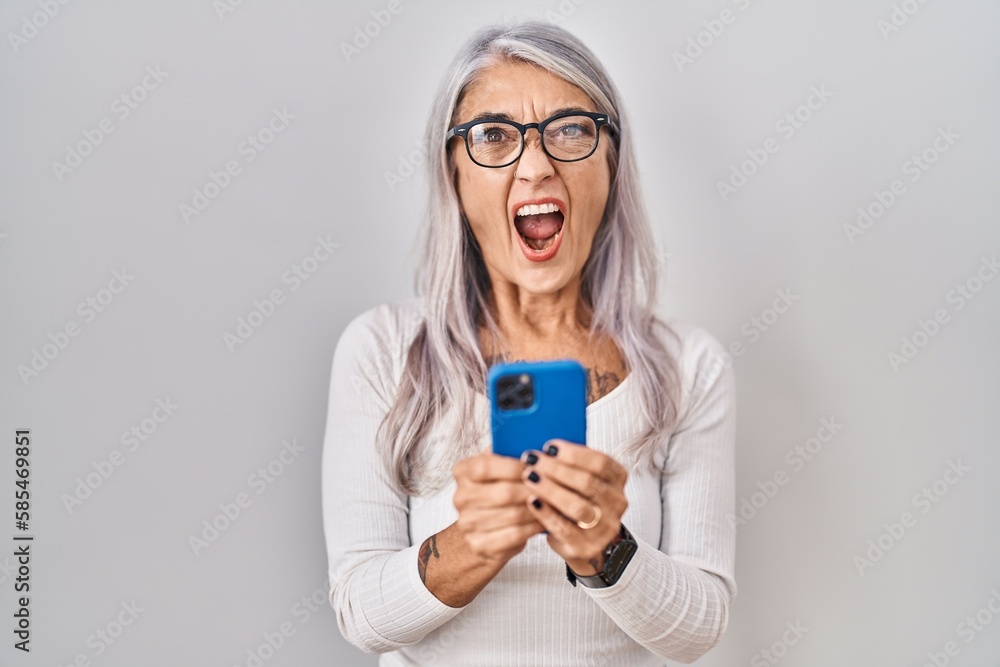 Middle age woman with grey hair using smartphone typing message angry and mad screaming frustrated and furious, shouting with anger. rage and aggressive concept. Middle age woman with grey hair using smartphone typing message angry and mad screaming frustrated and furious, shouting with anger. rage and aggressive concept.