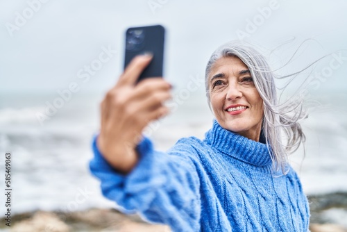 Middle age grey-haired woman smiling confident making selfie by the smartphone at seaside