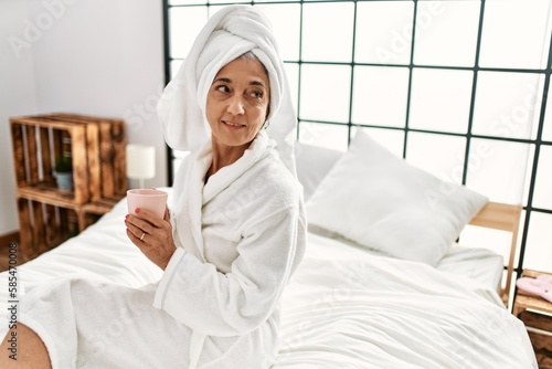 Middle age grey-haired woman drinking coffee sitting on bed at bedroom