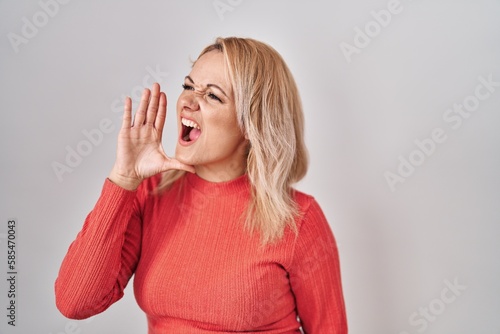 Blonde woman standing over isolated background shouting and screaming loud to side with hand on mouth. communication concept.