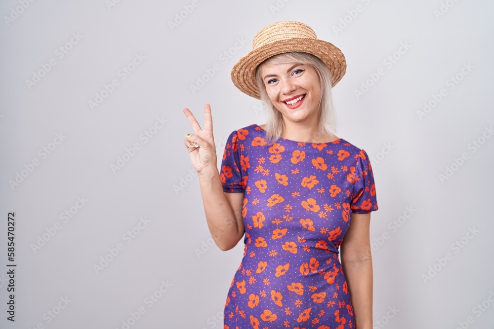 Young caucasian woman wearing flowers dress and summer hat showing and pointing up with fingers number two while smiling confident and happy. Young caucasian woman wearing flowers dress and summer hat showing and pointing up with fingers number two while smiling confident and happy.