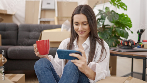 Young beautiful hispanic woman using smartphone drinking coffee sitting on floor at new home