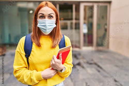 Young woman wearing safety mask and student backpack holding books scared and amazed with open mouth for surprise, disbelief face © Krakenimages.com