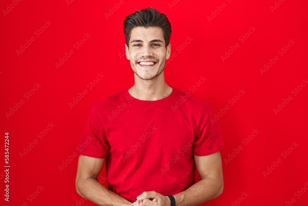 Young hispanic man standing over red background with hands together and crossed fingers smiling relaxed and cheerful. success and optimistic Young hispanic man standing over red background with hands together and crossed fingers smiling relaxed and cheerful. success and optimistic