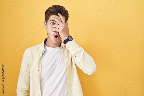 Young hispanic man standing over yellow background peeking in shock covering face and eyes with hand, looking through fingers with embarrassed expression.