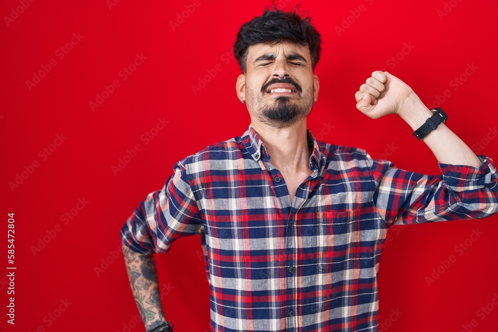 Fototapeta premium Young hispanic man with beard standing over red background stretching back, tired and relaxed, sleepy and yawning for early morning