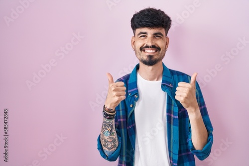 Young hispanic man with beard standing over pink background success sign doing positive gesture with hand, thumbs up smiling and happy. cheerful expression and winner gesture.