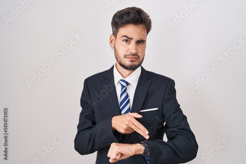 Young hispanic man with tattoos wearing business suit and tie in hurry pointing to watch time, impatience, upset and angry for deadline delay