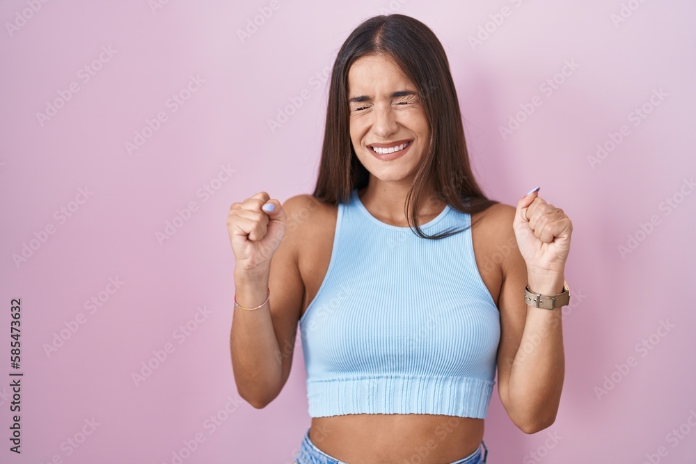 Young brunette woman standing over pink background excited for success with arms raised and eyes closed celebrating victory smiling. winner concept. Young brunette woman standing over pink background excited for success with arms raised and eyes closed celebrating victory smiling. winner concept.