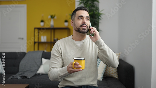Young hispanic man talking on smartphone drinking coffee at home