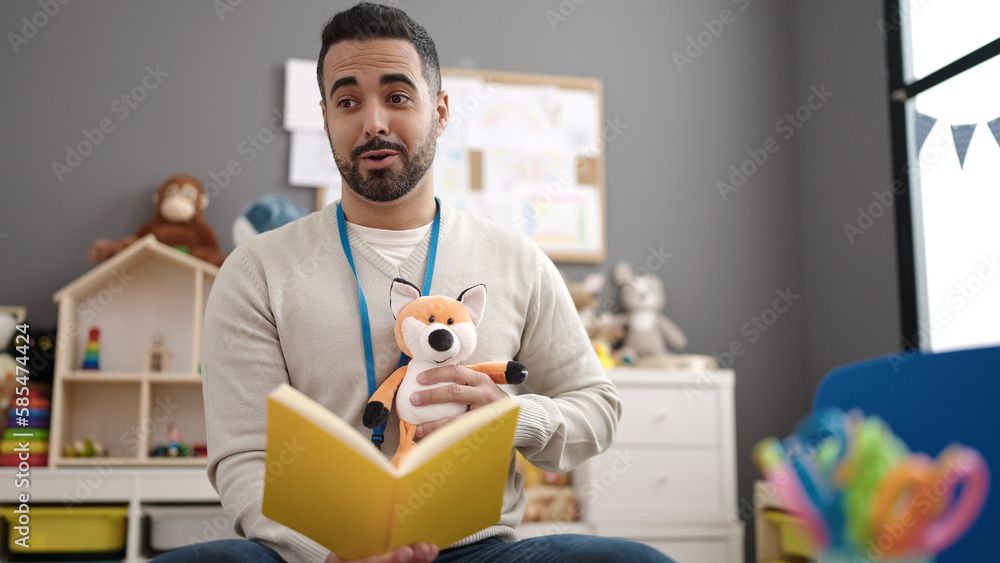 Young hispanic man preschool teacher reading story book sitting on table at kindergarten Young hispanic man preschool teacher reading story book sitting on table at kindergarten