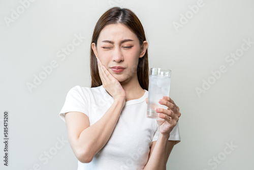 Suffering from toothache, blur asian young woman touch cheek, face expression ache or feel pain, sensitive molar teeth, hand holding glass of water with ice, inflammation when drink cold, healthcare.