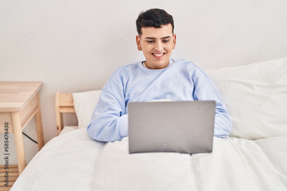Young non binary man using laptop sitting on bed