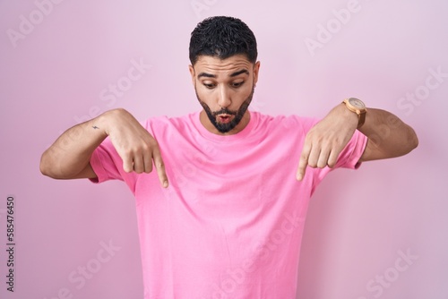 Hispanic young man standing over pink background pointing down with fingers showing advertisement, surprised face and open mouth