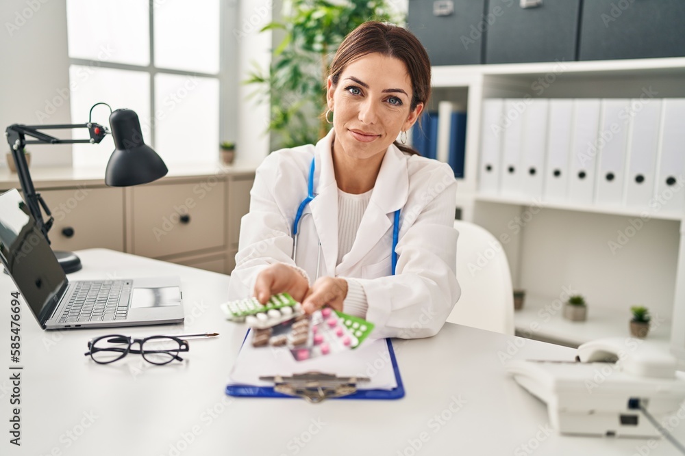 Young brunette doctor woman holding pills smiling with a happy and cool smile on face. showing teeth. Young brunette doctor woman holding pills smiling with a happy and cool smile on face. showing teeth.