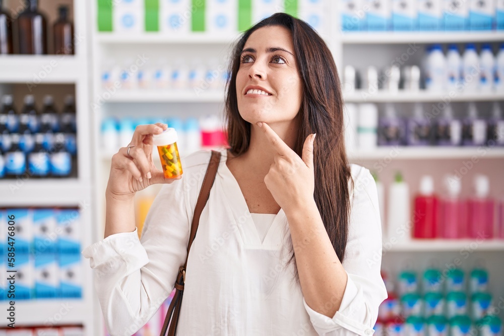 Young brunette woman shopping at pharmacy drugstore holding pills serious face thinking about question with hand on chin, thoughtful about confusing idea