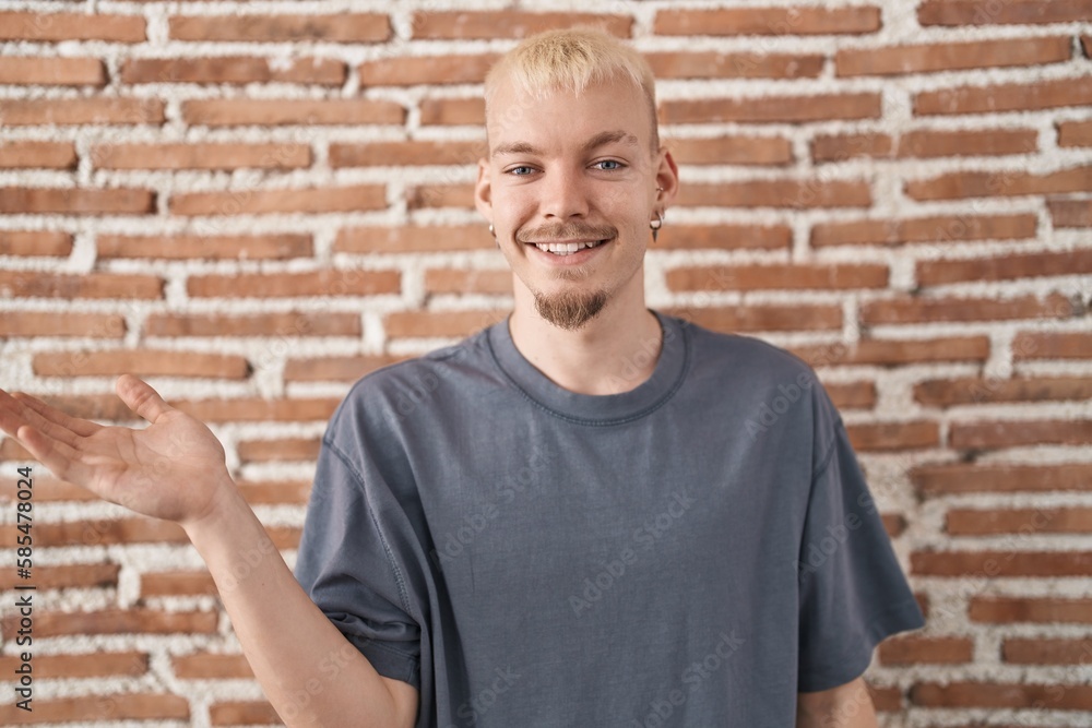 Young caucasian man standing over bricks wall smiling cheerful presenting and pointing with palm of hand looking at the camera. Young caucasian man standing over bricks wall smiling cheerful presenting and pointing with palm of hand looking at the camera.
