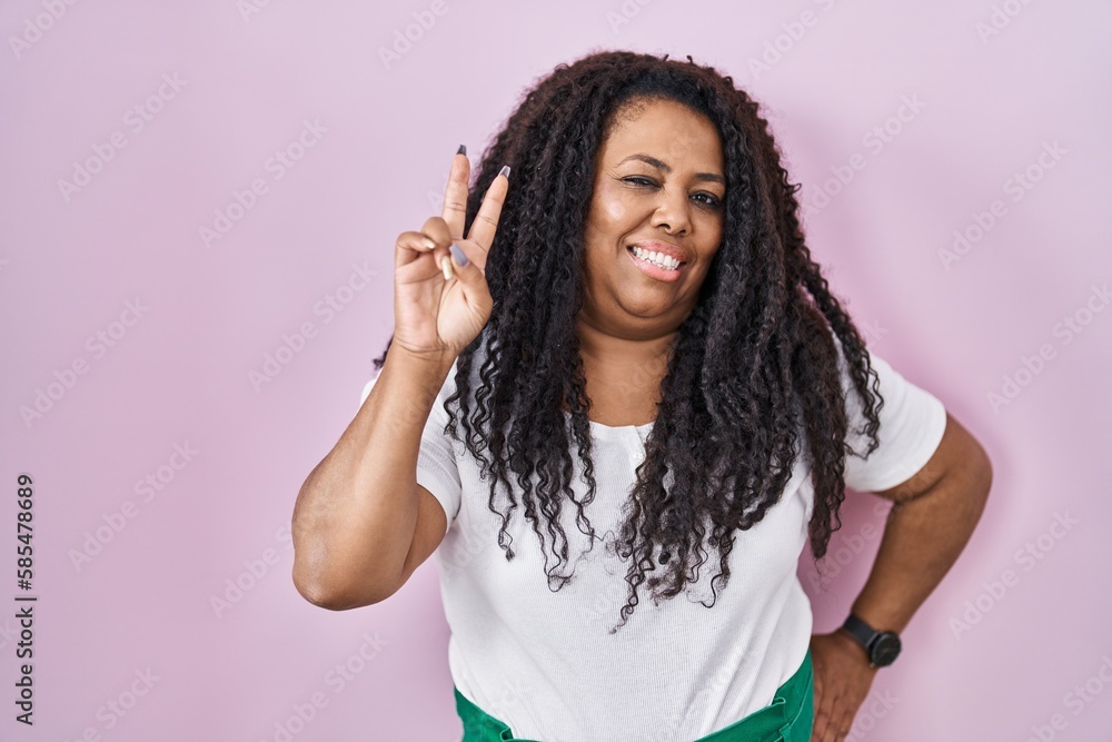 Plus size hispanic woman standing over pink background smiling looking to the camera showing fingers doing victory sign. number two. Plus size hispanic woman standing over pink background smiling looking to the camera showing fingers doing victory sign. number two.