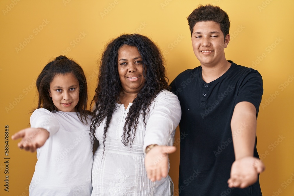 Family of mother, daughter and son standing over yellow background smiling cheerful offering palm hand giving assistance and acceptance. Family of mother, daughter and son standing over yellow background smiling cheerful offering palm hand giving assistance and acceptance.