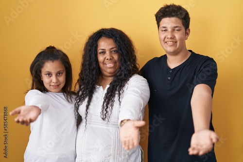 Family of mother, daughter and son standing over yellow background smiling cheerful offering palm hand giving assistance and acceptance.