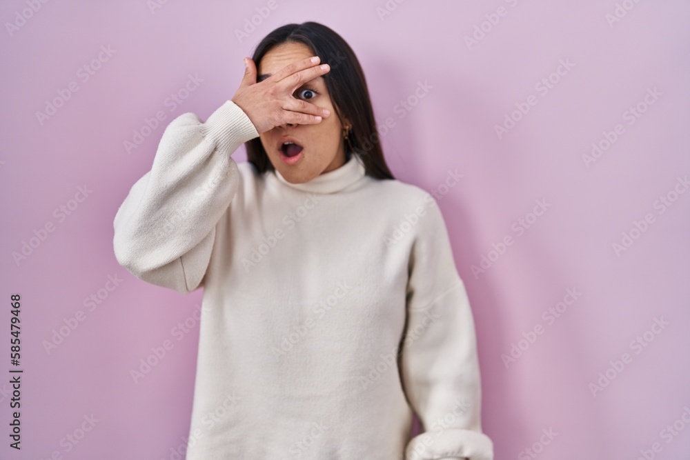 Young south asian woman standing over pink background peeking in shock covering face and eyes with hand, looking through fingers with embarrassed expression. Young south asian woman standing over pink background peeking in shock covering face and eyes with hand, looking through fingers with embarrassed expression.