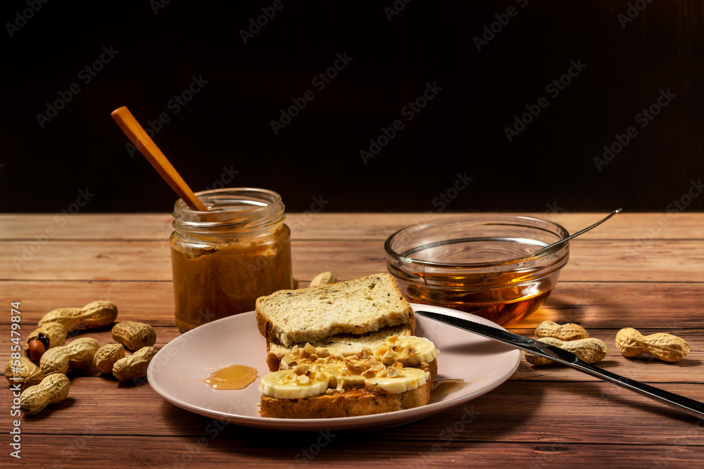 Peanut butter or peanut paste in an open jar and spread on a toast sandwich with banana and honey and peanuts in shells scattered on the table